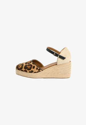 Piedi che indossano sandali espadrillas con zeppa, con parte anteriore stampa leopardata, tacco beige intrecciato e cinturini neri alla caviglia con fibbie su un tappeto crema.