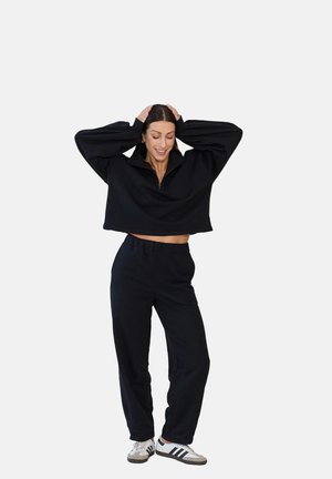 Kobieta uśmiecha się, trzymając ręce na głowie, ubrana w czarną krótką bluzę zapinaną na zamek, czarne spodnie dresowe oraz białe sneakersy z czarnymi paskami.