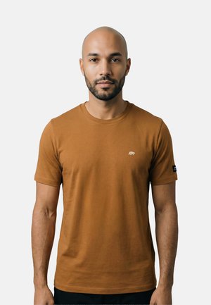 Mann mit kurzem Bart, der ein schlichtes braunes T-Shirt mit kurzen Ärmeln trägt, auf der Brust ein kleines Bärenlogo und auf dem linken Ärmel ein Aufnäher.