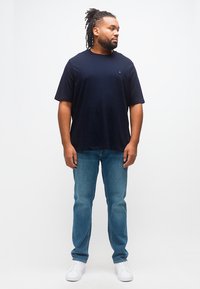 Άνδρας με ντραντλοκς, ντυμένος με μαύρο μπλουζάκι, μπλε τζιν και άσπρα αθλητικά παπούτσια, στέκεται και κοιτάζει προς το πλάι μπροστά σε απλό φόντο.