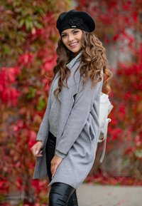 Šedý oversized kardigan s texturovaným svetrém pod ním, černý baret a černé kalhoty, doplněné bílým batohem. Červené listí v pozadí.