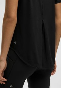 Zwarte sportieve shirt met een licht gebogen onderkant, voorzien van een ventilerend detail aan de achterkant. Draagt met zwarte leggings, waar het logo nabij de naad zichtbaar is.
