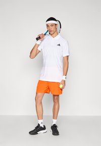 Λευκό κοντομάνικο πουκάμισο πόλο με κάθετες ρίγες, πορτοκαλί σορτς, μαύρα παπούτσια τένις και ρακέτα τένις. Κρατάει κίτρινη μπάλα τένις.