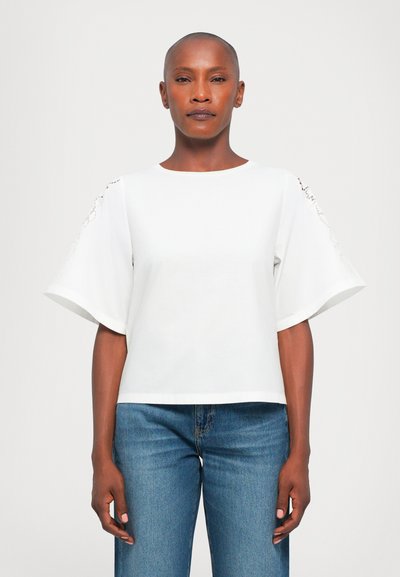 Haut blanc à manches courtes avec une coupe ample et un col rond. Présente des détails en dentelle sur les manches. Associé à un jean en denim bleu.
