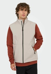 Beige ärmellose Weste mit hohem Kragen und Reißverschluss, ausgestattet mit roten langen Ärmeln, Seitentaschen und schwarzem Reißverschluss-Detail.
