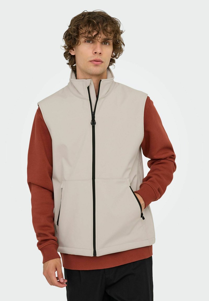 Beige ärmellose Weste mit hohem Kragen und Reißverschluss, ausgestattet mit roten langen Ärmeln, Seitentaschen und schwarzem Reißverschluss-Detail.