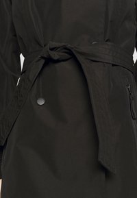 Primo piano di un cappotto nero legato in vita con una cintura cucita, rifinito con un bottone a pressione e una tasca con zip.