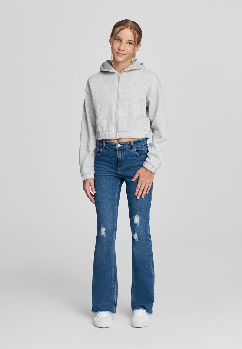 Felpa grigia cropped con zip frontale e tasche, abbinata a jeans a zampa d'elefante blu con dettagli strappati e sneakers bianche.