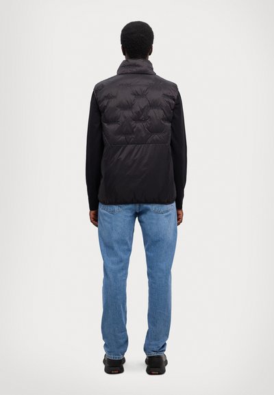 Μαύρο γιλέκο πούφ με ψηλό γιακά, με υφάσματα σε καπιτονέ σχέδιο και ελαφρύ υλικό, συνδυασμένο με ανοιχτόχρωμα τζιν και σκούρα παπούτσια.