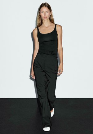 Černý top na ramínka a kalhoty s vysokým pasem z hladkého materiálu. Model má na sobě bílé slip-on boty. Minimalistický design na jednobarevném pozadí.