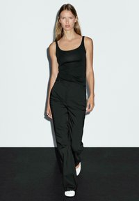 Schwarzes, tailliertes Tanktop kombiniert mit weiten schwarzen Hosen. Weiße Schuhe runden den Look ab. Glattes Material mit minimalistischem Design.