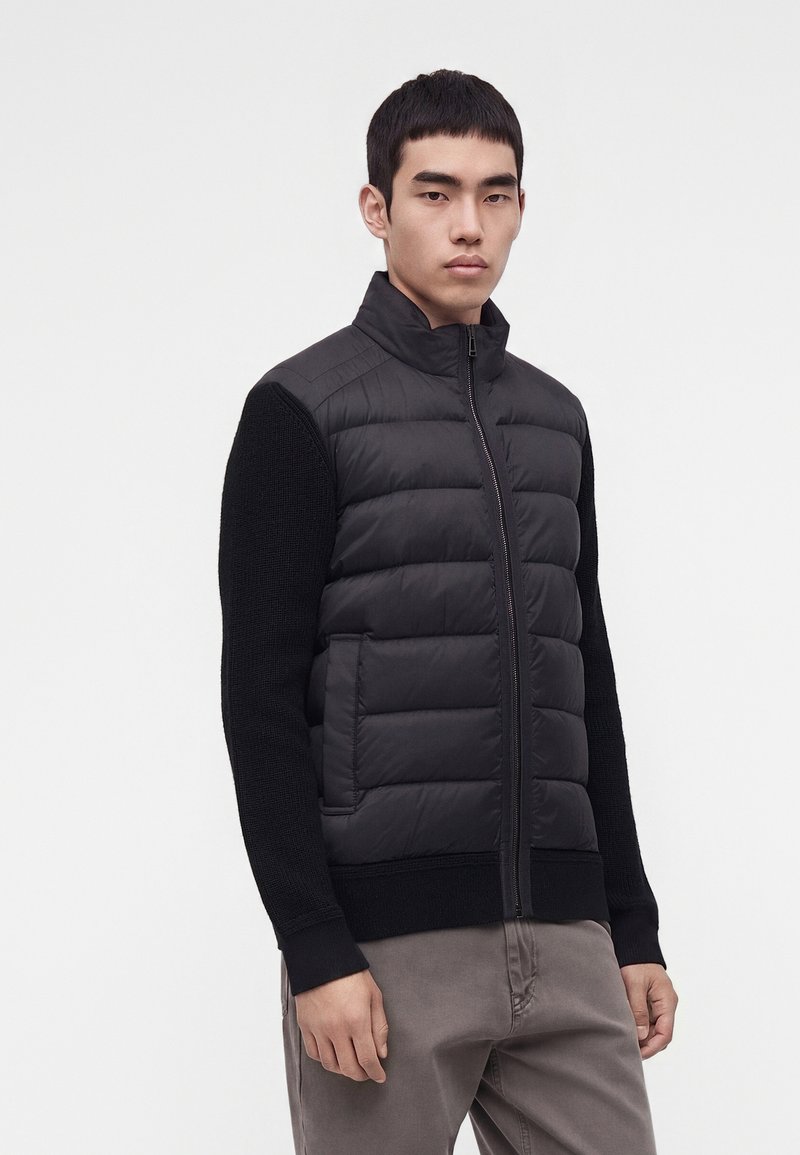 Jeune homme portant une veste matelassée noire zippée avec des manches en tricot et un pantalon gris, debout devant un fond clair uni.