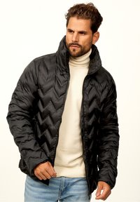 Black puffer jacket with a chevron quilted pattern, high collar, and zip closure. Worn over a beige turtleneck sweater and blue jeans.