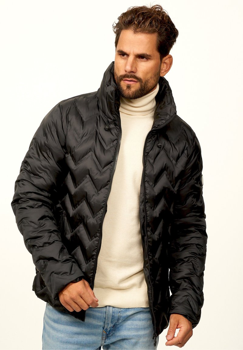 Black puffer jacket with a chevron quilted pattern, high collar, and zip closure. Worn over a beige turtleneck sweater and blue jeans.