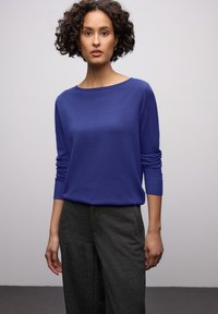 Pull tricot bleu roi avec un col bateau et des manches longues, associé à un pantalon ample gris foncé. Texture lisse, coupe décontractée.