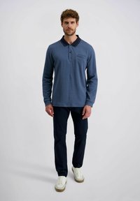 Mann steht und blickt nach vorne, trägt ein blaues Langarm-Polohemd, dunkle Hosen und weiße Sneaker vor einem schlichten, hellen Hintergrund.