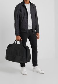 Svart rutig jacka, grå tröja, svarta jeans och vita sneakers. Håller en svart träningsväska med matt finish och robusta handtag.