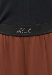 Fustă de culoare ruginită, cu pliuri, având un talie neagră. Talia are un logo brodat "Karl" cu fir argintiu.