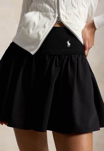 Kvinne iført svart skjørt med vide slag og elastisk midje med hvit Polo Player-logo, kombinert med en hvit kabelstrikket genser med glidelås.