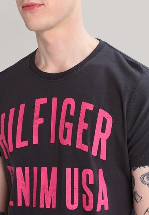 Camiseta de algodón negra con un atrevido estampado rosa "HILFIGER DENIM USA". Mangas cortas, cuello redondo y textura suave con sutiles detalles de costura.