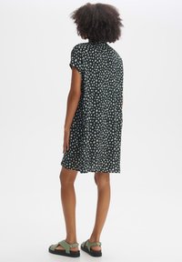 Femme aux cheveux bouclés portant une petite robe noire à pois blancs et des sandales vertes, debout de dos sur un fond blanc.