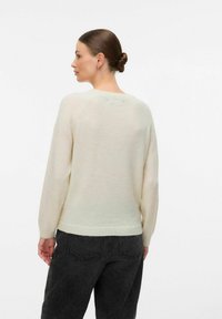 Mujer con el cabello recogido en un moño, que lleva pequeños pendientes en forma de aro, un suéter de punto color crema y jeans negros, de espaldas contra un fondo blanco simple.