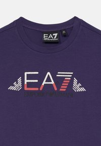 Mörklila t-shirt med en skinande silver- och röd "EA7 Emporio Armani" logotyp tryckt på bröstet under en svart märkeetikett vid halslinningen.