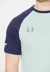Lysgrøn og marineblå sports skjorte med korte ærmer, med et logo af en ræv og kontrasterende sømdetaljer. Glat tekstur.