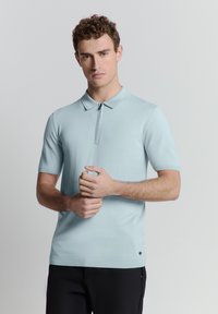 Lichtblauw polo shirt met korte mouwen en een ritskraag, van gestructureerde stof en subtiele logodetail aan de onderrand. Gestyled met donkere broek.