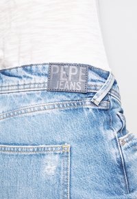 Gaiši zili džinsi no denima, kuriem ir ādas uzlīme ar iegravētu "PEPE JEANS". Iekļauti šuvju detaļas un aizmugurējais kabata ar saplaisājušām malām.