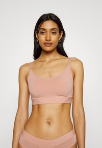Light pink bralette made of stretchy fabric, featuring a rounded neckline and adjustable shoulder straps. Smooth texture, minimal design.