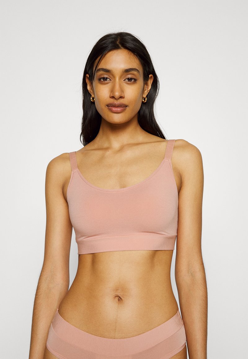 Light pink bralette made of stretchy fabric, featuring a rounded neckline and adjustable shoulder straps. Smooth texture, minimal design.