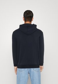 Felpa blu navy realizzata in tessuto morbido con un ampio cappuccio, polsini e orlo a coste. Indossata con pantaloni di denim azzurri, vista da dietro.