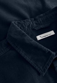 Col de chemise en velours côtelé bleu foncé avec une étiquette blanche visible montrant un logo de hibou et le texte "Knowledge Cotton Apparel".