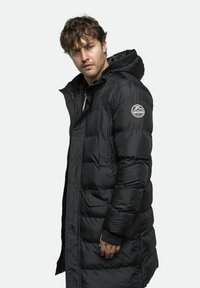 Dug crni puffer kaput s kapuljačom, napravljen od sjajnog materijala. Ima rebraste manžetne, bočne džepove i logotip na lijevoj ruci.