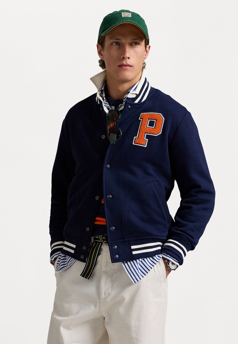 Námornícky varsity kabát s oranžovým logom "P", bielymi pruhovanými rukávmi a cvokmi; vrstvený cez pruhované tričko s zelenou čiapkou.