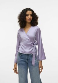 Haut enveloppant en satin lilas à manches longues, avec un décolleté en dentelle et un accent en nœud à la taille, associé à un jean bleu clair.