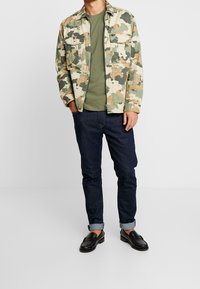 Giacca a motivo camouflage in beige, verde e marrone, sovrapposta a una t-shirt verde oliva; jeans scuri e mocassini neri, con i polsini arrotolati visibili.