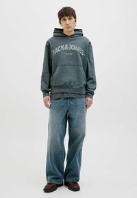 Zelena dukserica s logotipom "JACK&JONES", s kenguruljom, u kombinaciji s svjetloplavim širokim trapericama i smešnim cipelama. Jednostavan dizajn i opušteni kroj.