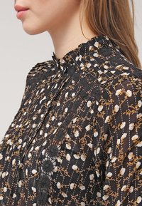 Blouse noire à motif floral en blanc et or, avec des accents en dentelle, un col montant et un design plissé. Tissu semi-transparent.