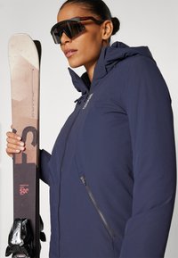 Navy insulated ski jacket with a high collar, adjustable hood, and a side zipper pocket, paired with black ski goggles and a patterned ski.