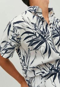 Camicia a maniche corte in tessuto leggero, con sfondo bianco e stampe di foglie tropicali nere, colletto con bottoni.