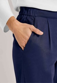Marineblauwe broek met een geplooide ontwerp, voorzien van een elastische tailleband. De hand ligt in een zijzak, wat de gladde stoftextuur laat zien.