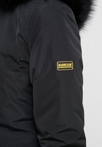 Veste noire isolante avec capuche en fausse fourrure et logo Barbour International orné de détails dorés sur la manche gauche. Texture lisse.