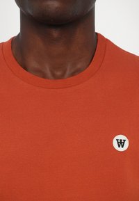 Sarkana kokvilnas T-krekls ar apaļu kakla izgriezumu un mazu balto izņemta logotipu kreisajā pusē. Gluda tekstūra, vienkāršs dizains.