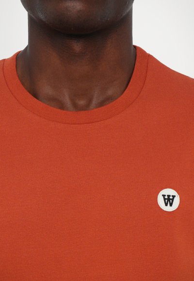 Red cotton T-shirt with a round neckline and small white embroidered logo on the left side. Smooth texture, simple design.