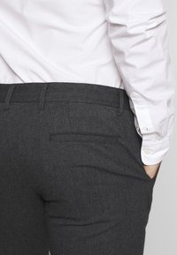 Muž oblečený v tmavě šedých kalhotách s proužky a bílém tričku s dlouhým rukávem, pravá ruka v kapse, zobrazený od pasu dolů.