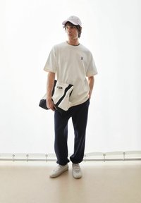 Wit T-shirt met een paars lintlogo, donkerblauwe broek en een roze pet. Houdt een tweekleurige zwarte en crèmekleurige tas met een logo vast. Witte schoenen.