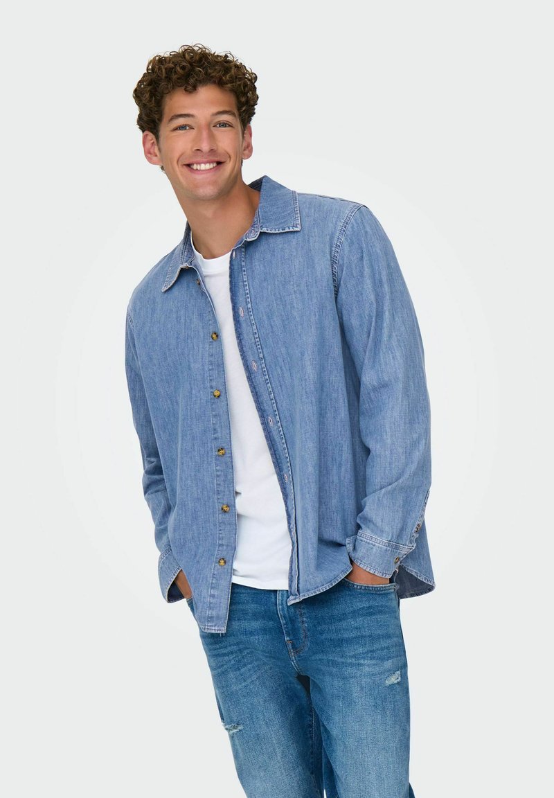 Lichtblauwe denim blouse met lange mouwen, ontspannen pasvorm en gouden knopen, gedragen over een wit T-shirt, gecombineerd met blauwe jeans.