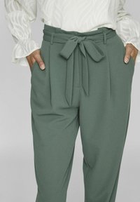 Green high-waisted trousers with a tied belt, pleats, side pockets, and a smooth texture. Paired with a white, textured blouse.